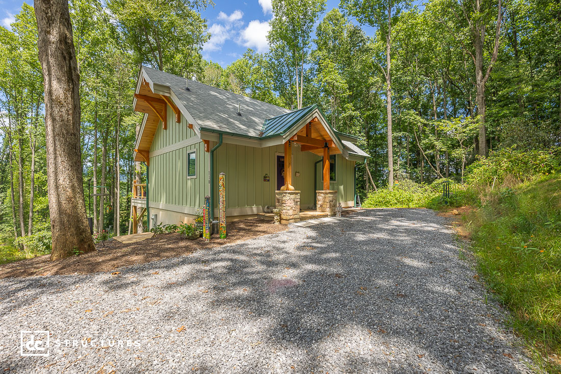 A small green house with a gabled roof and stone pillars in a wooded area, surrounded by trees and a gravel driveway under blue sky.