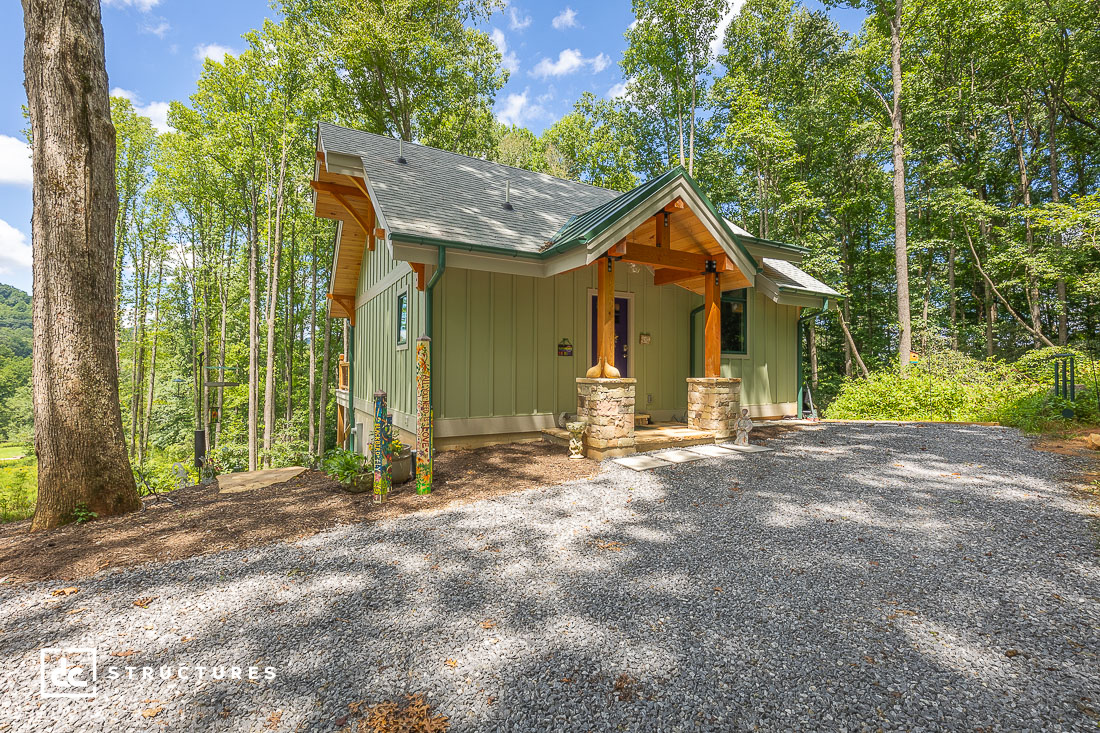 A small green cabin with a pitched roof, stone columns, and wooden trim sits in the woods on a gravel driveway under a blue sky.