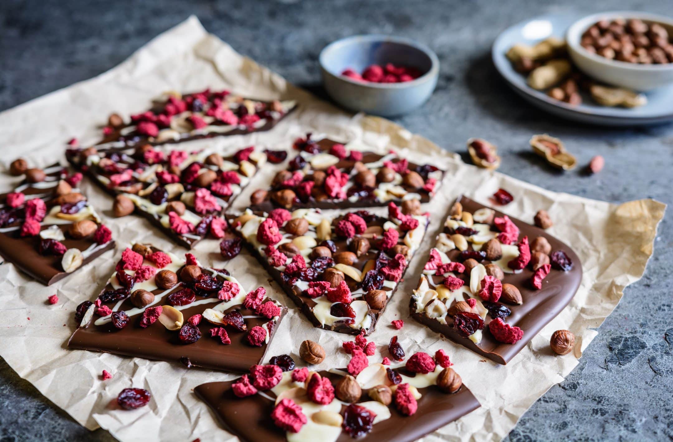 Chocolate bark pieces topped with dried raspberries, cranberries, nuts, and seeds on parchment paper, bowls of nuts and raspberries behind.