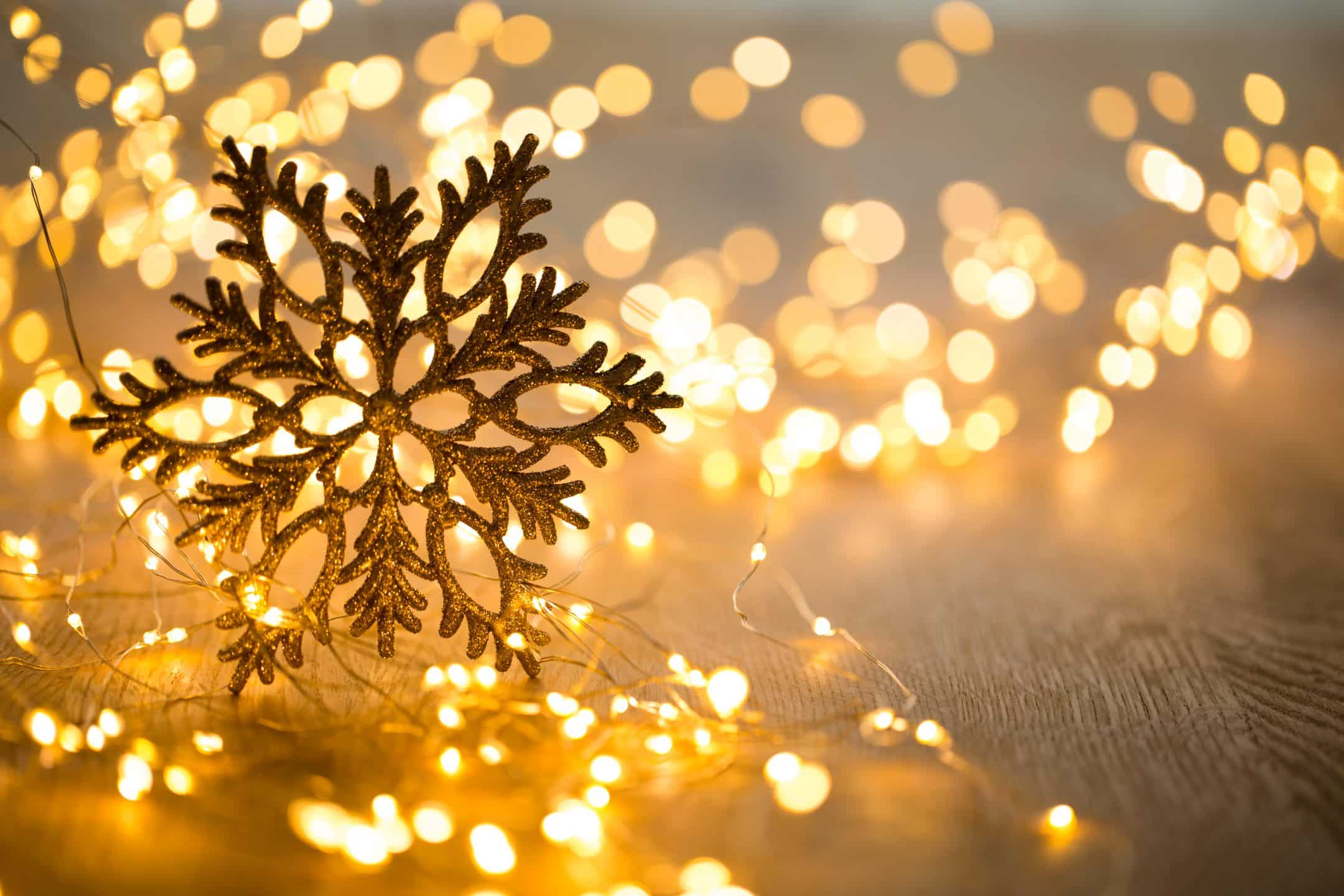 A glittery gold snowflake ornament stands among glowing yellow fairy lights on a wooden surface, with blurred festive background.
