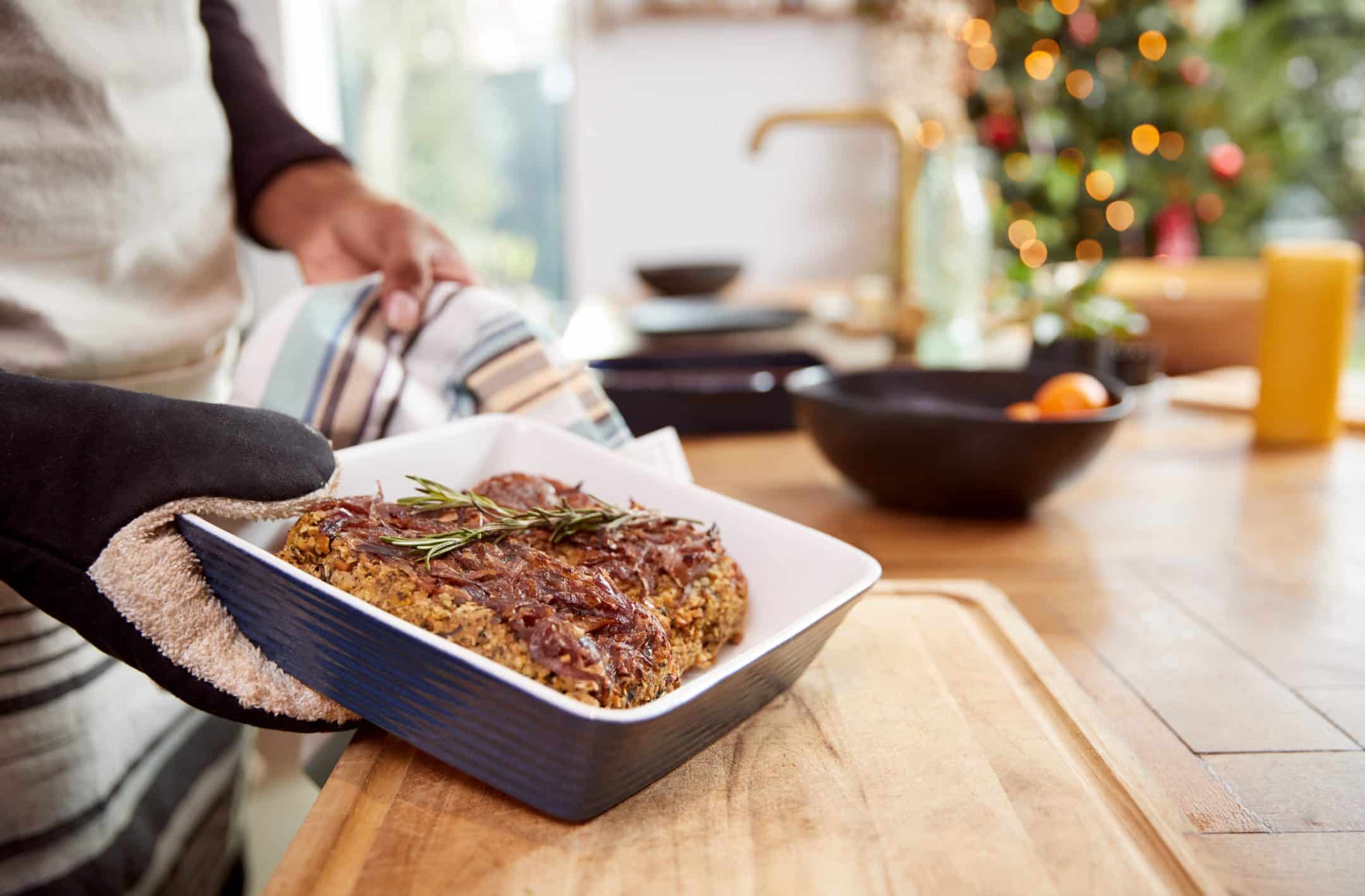 A person with an oven mitt sets a baking dish of herb-topped meatloaf on a wooden counter, Christmas tree in the background.