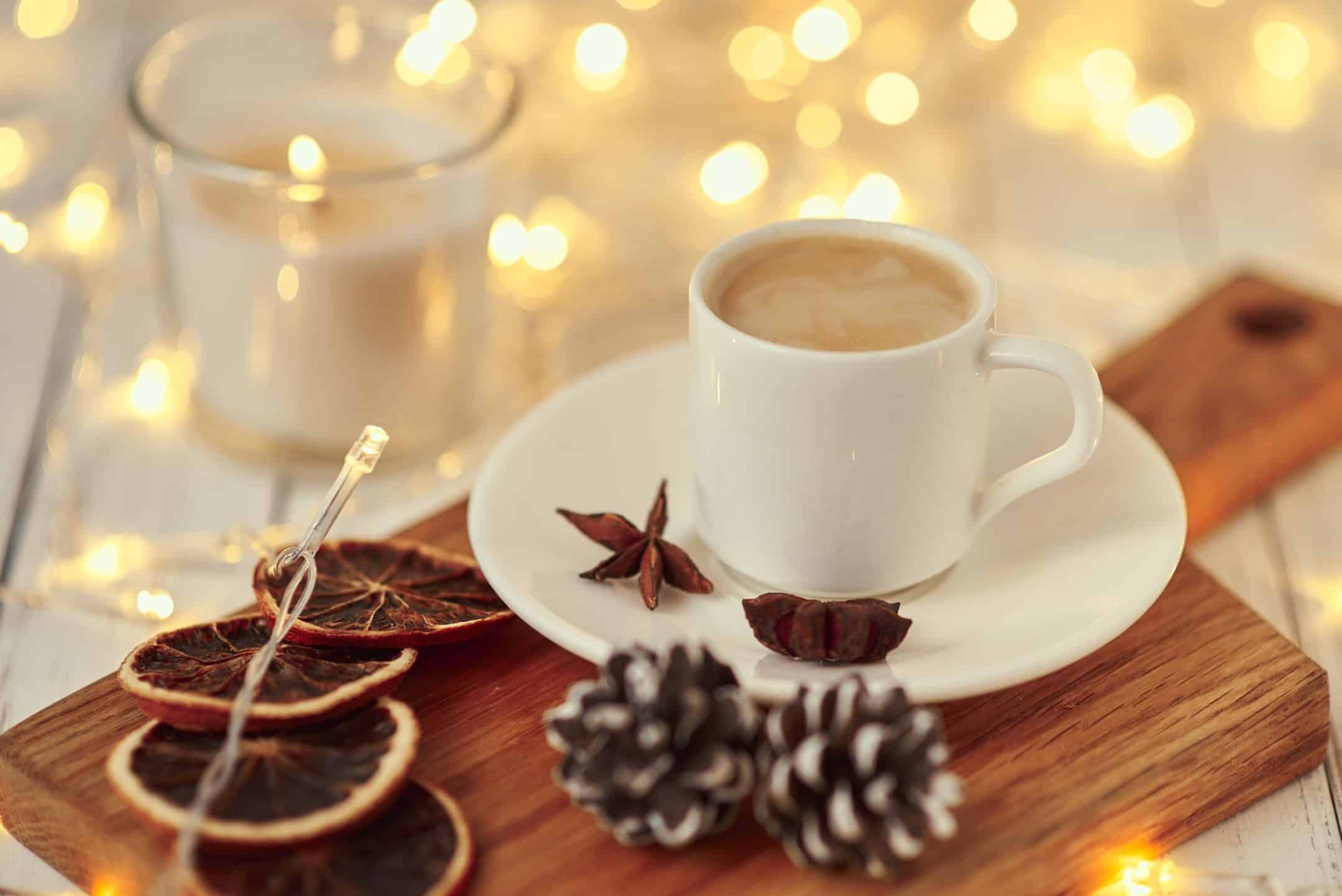 A white cup of coffee on a saucer with star anise, next to pinecones, dried oranges, a lit candle, and fairy lights in the background.
