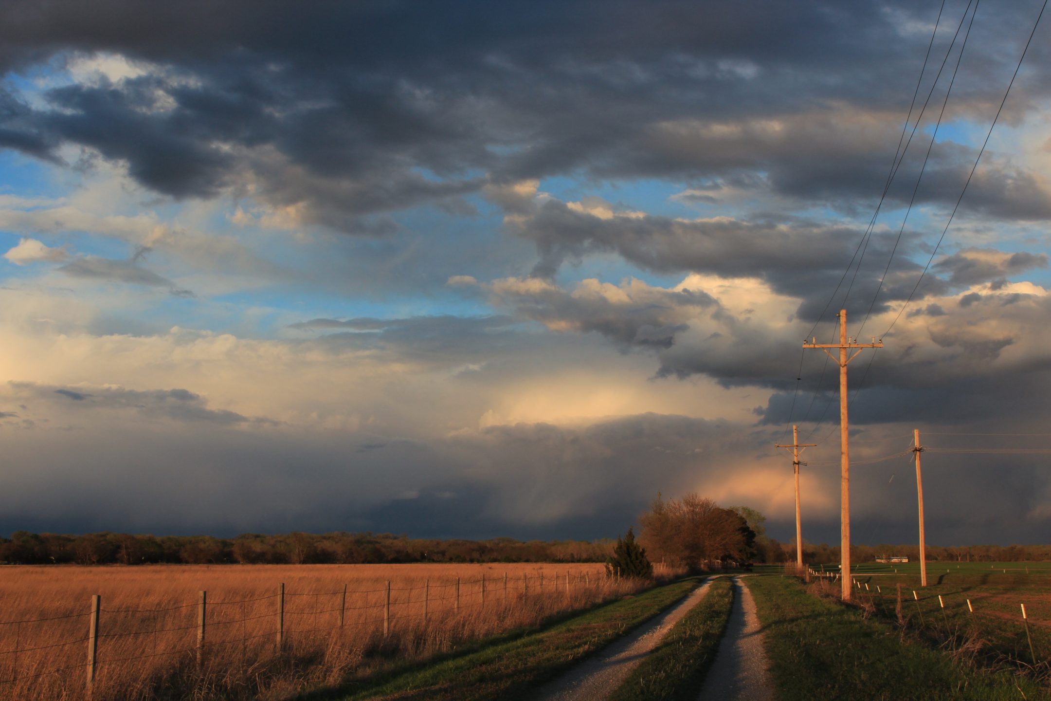 http://A%20rural%20dirt%20road%20runs%20alongside%20utility%20poles%20and%20fences,%20bordered%20by%20open%20fields%20under%20a%20dramatic%20sky%20with%20dark%20clouds%20and%20sunlight.