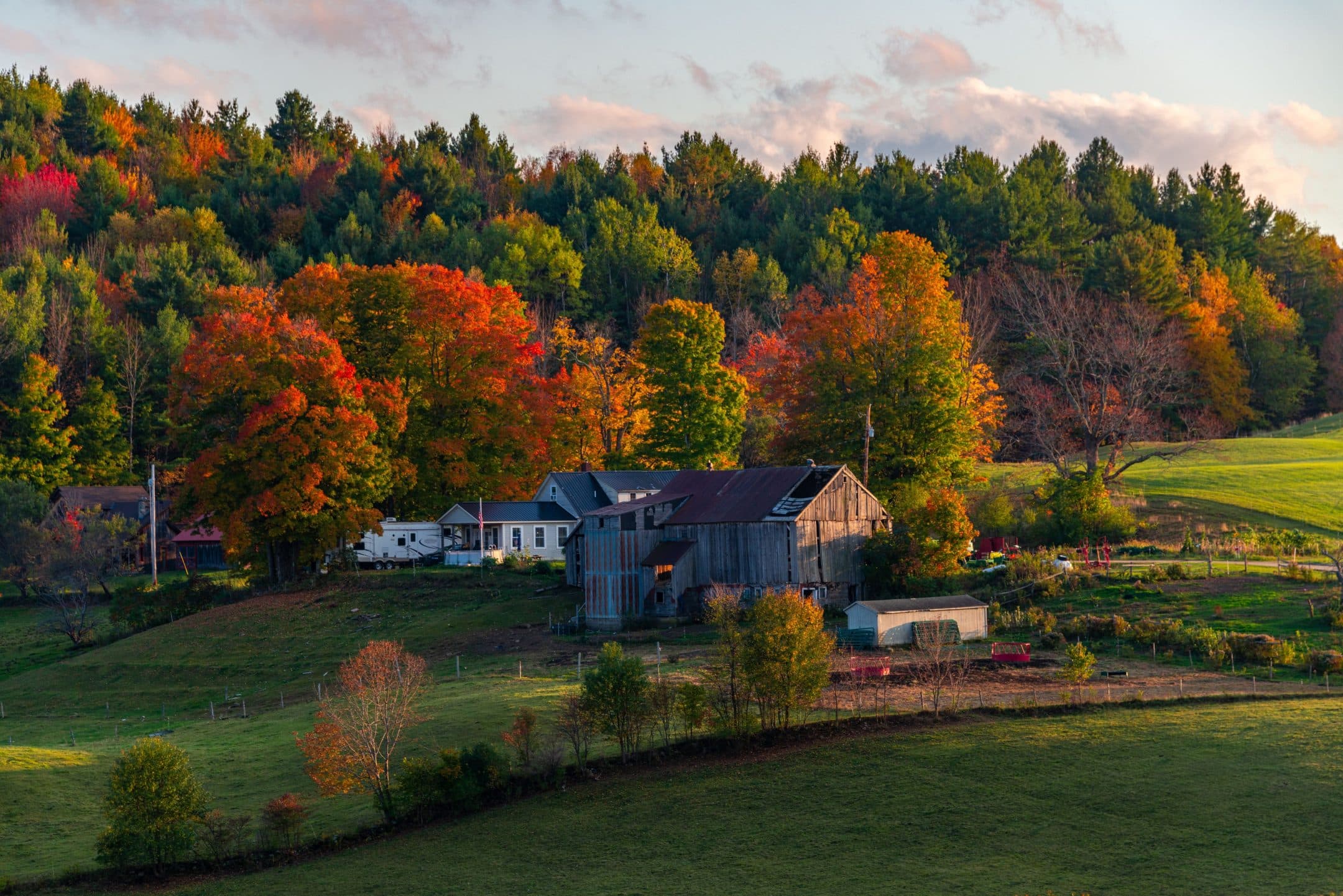 http://A%20rustic%20farmhouse%20and%20barn%20among%20vibrant%20autumn%20trees%20at%20a%20forest's%20edge,%20with%20colorful%20leaves%20and%20a%20grassy%20field%20below%20a%20cloudy%20sky.