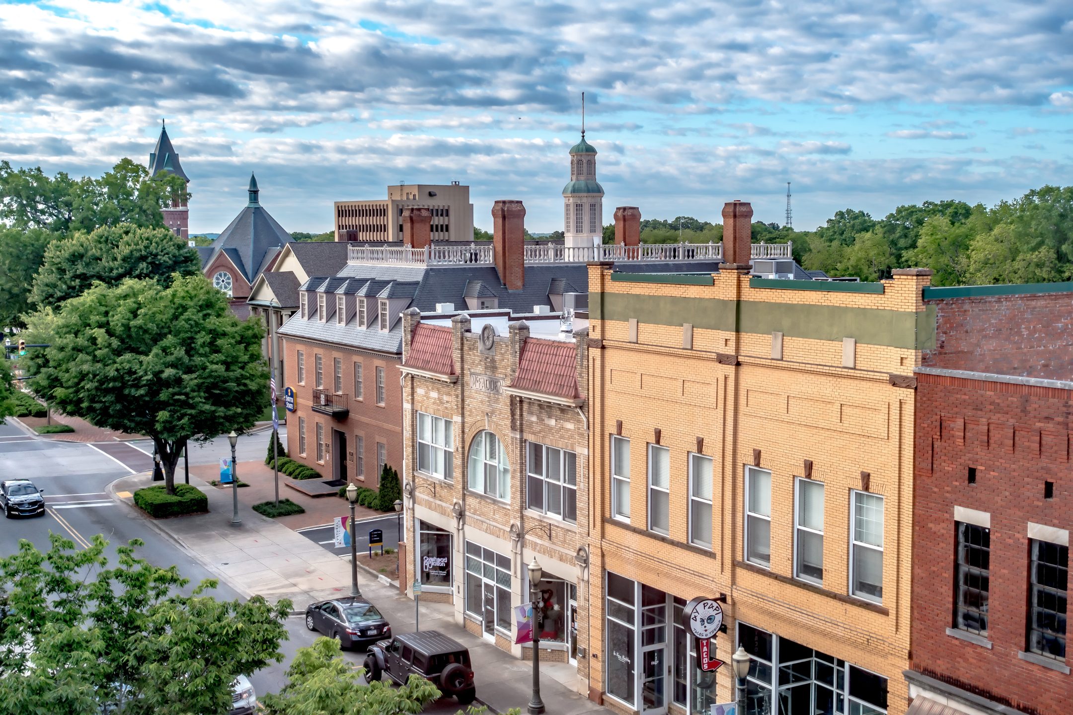 http://A%20vibrant%20small-town%20street%20with%20historic%20brick%20buildings,%20storefronts,%20parked%20cars,%20lush%20trees,%20and%20a%20church%20steeple%20in%20back.