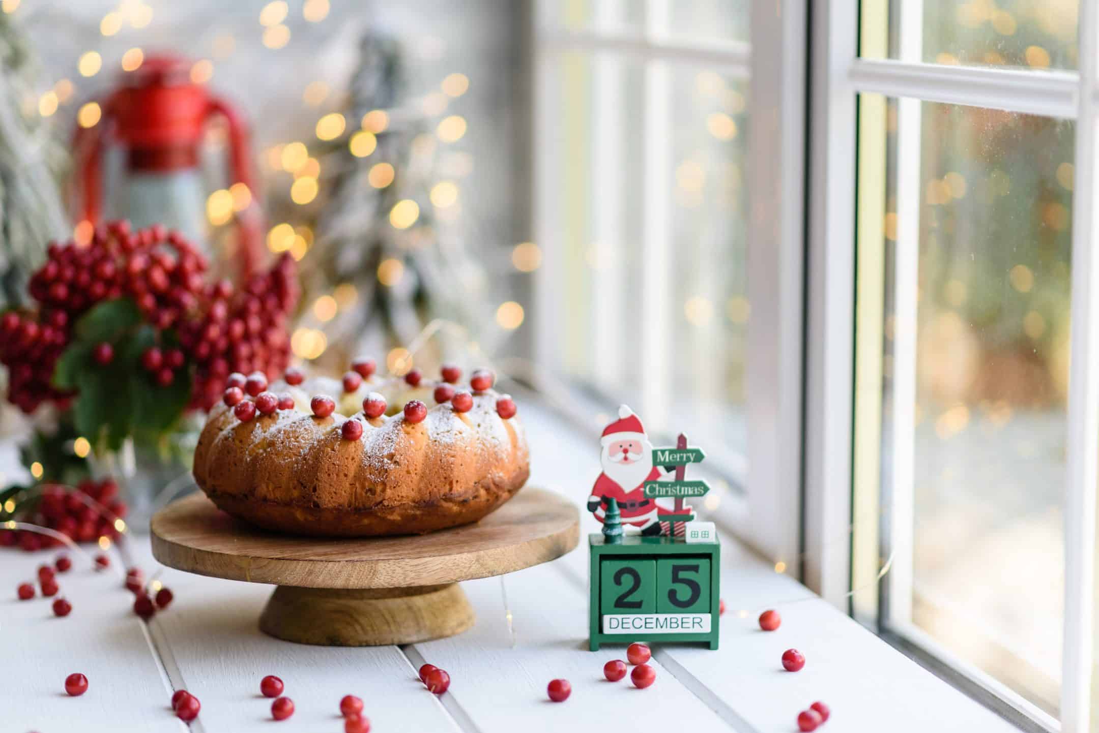 A festive bundt cake with cranberries sits on a wooden stand. Fairy lights, a red teapot, berries, and a snowy window create a cozy holiday scene.