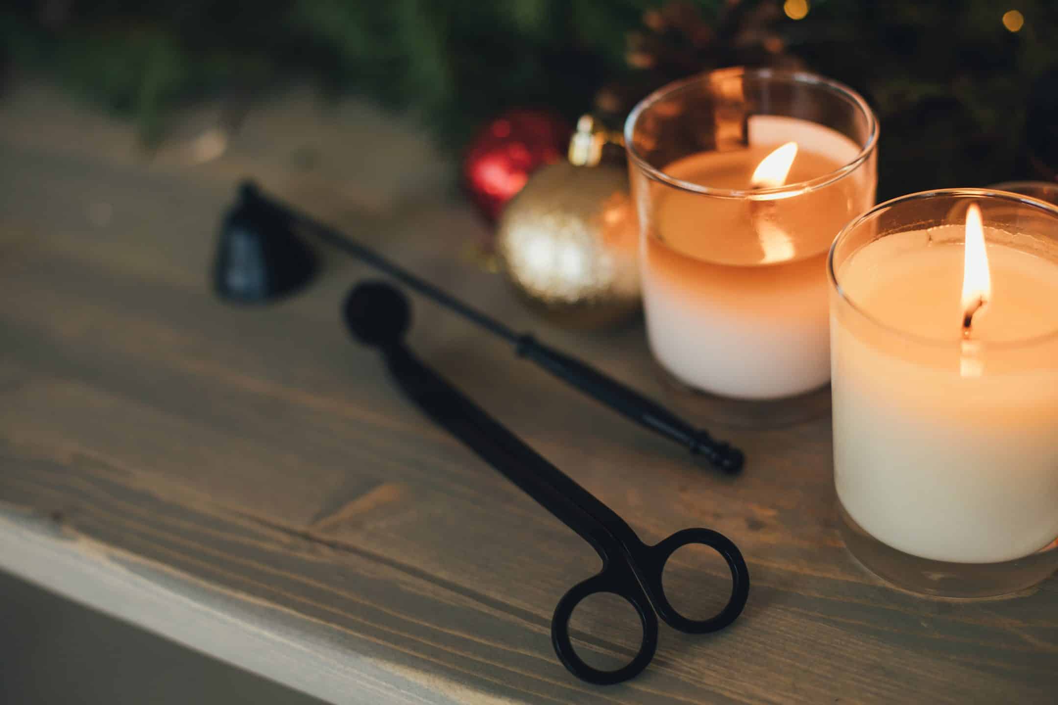 Two lit candles sit on a wooden surface beside a candle snuffer and wick trimmer, with blurred Christmas ornaments and greenery in the background.
