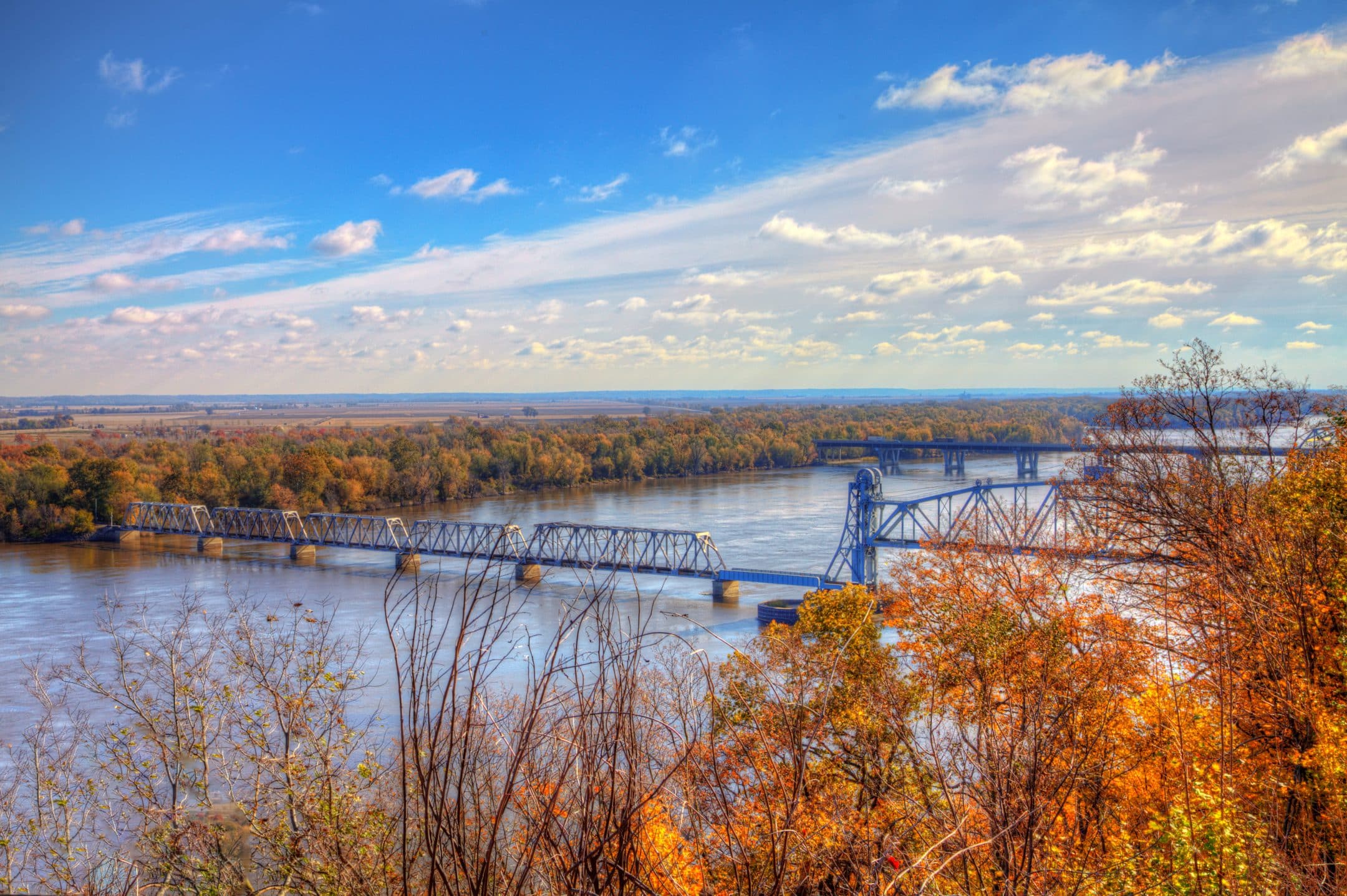 http://A%20steel%20railroad%20bridge%20spans%20a%20wide%20river%20surrounded%20by%20orange%20and%20yellow%20autumn%20trees,%20with%20a%20vast%20landscape%20and%20blue%20sky%20behind.