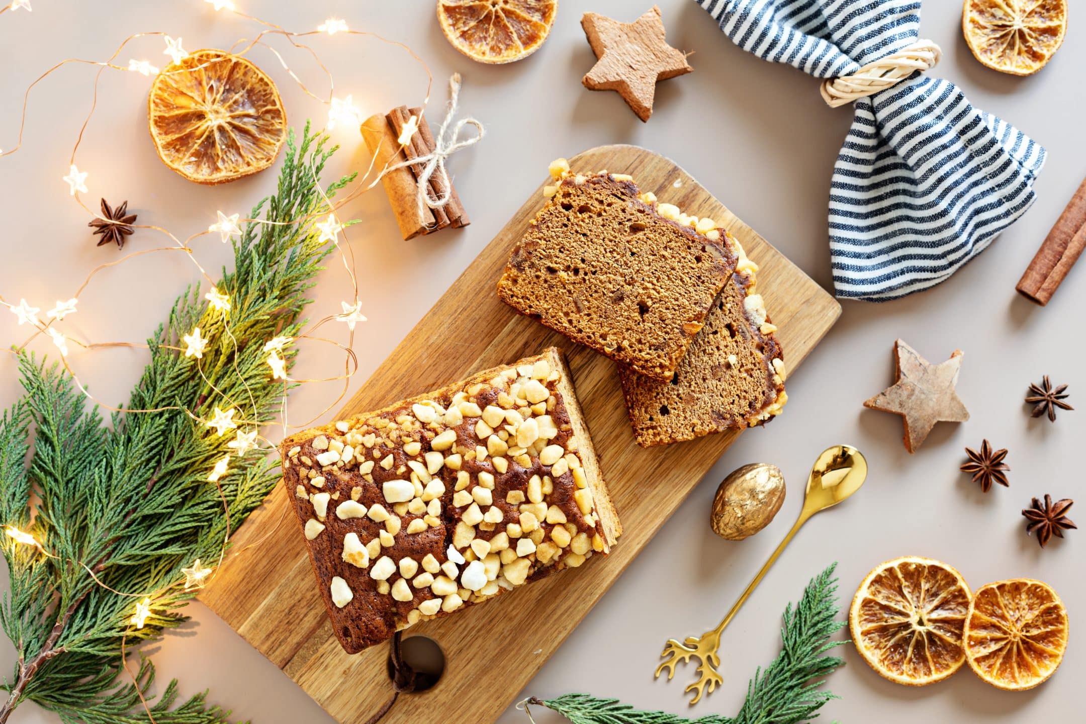 A loaf of nut-topped cake, partially sliced, on a wooden board with festive decor like dried orange slices, spices, and string lights.