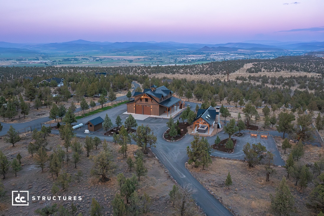 Aerial view of two large rustic buildings surrounded by trees and winding roads in a rural landscape at sunset, with mountains and a valley visible in the distance.