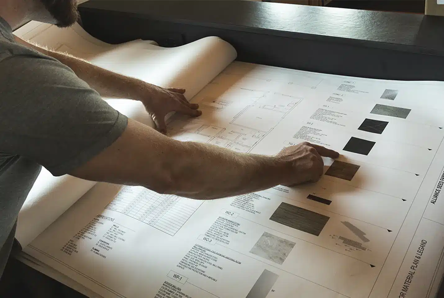 A person points to material samples and specifications on a large architectural blueprint or design plan spread across a table.