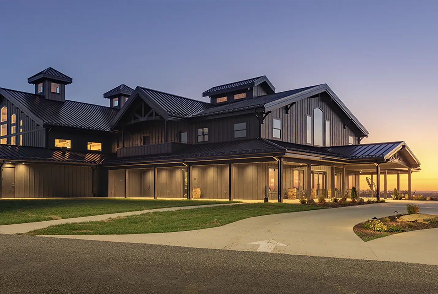 A large modern barn-style building with dark metal siding and a metal roof is warmly lit at sunset, with gables, tall windows, and a curved driveway on a green lawn.