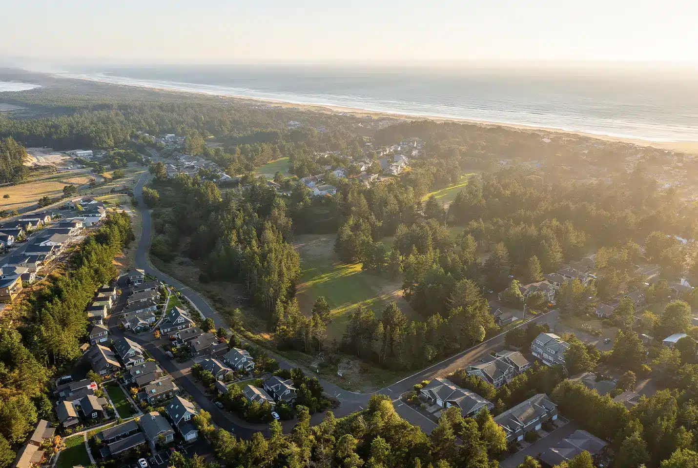 Aerial view of a coastal town with rows of houses, dense green trees, and a long sandy beach along the ocean in golden sunlight.