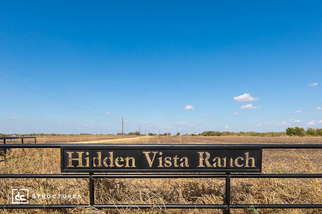 A metal gate sign in front of a wide, open field under a clear blue sky with a few clouds. Power lines and distant trees are visible.