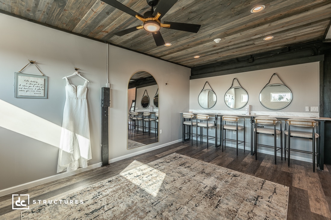 A modern rustic room with wood floors and ceiling, a hanging white dress, arched and round mirrors, stools, and sunlight streaming in.