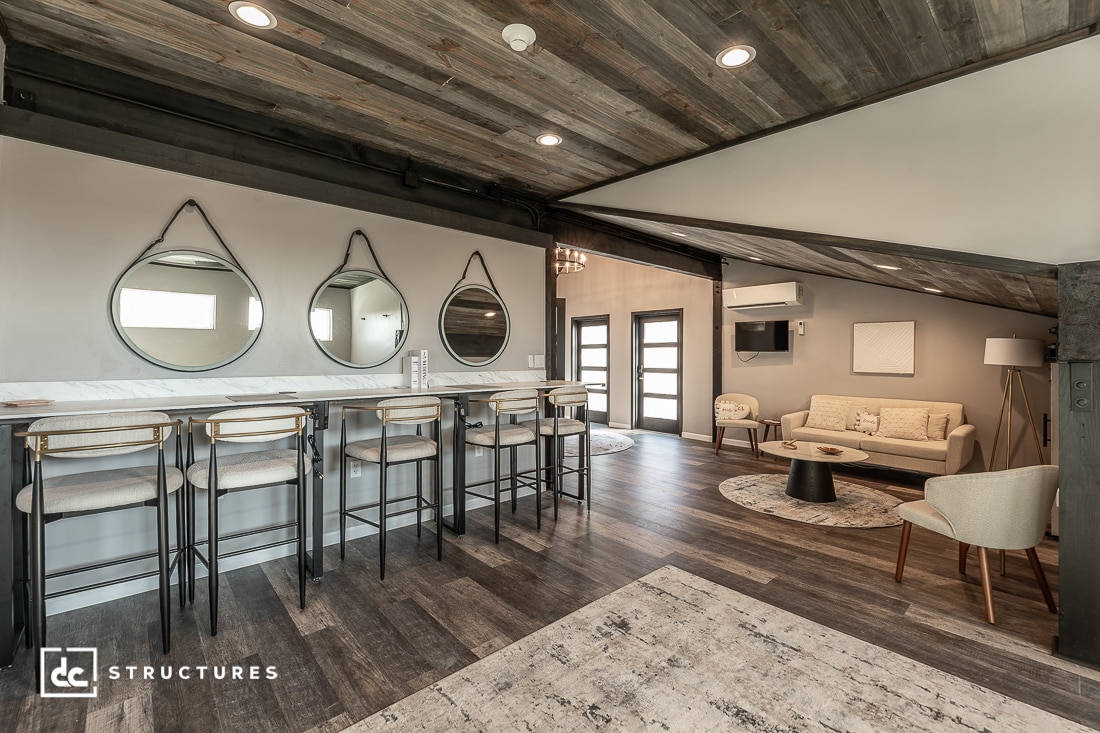 A stylish open-concept loft with a marble bar, four stools, three round mirrors, a cozy sitting area, wood floors, and a rustic sloped ceiling.
