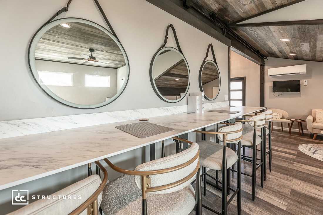 A modern workspace with a long marble counter, stylish bar stools, three round mirrors, wood-accented ceilings, and natural lighting.