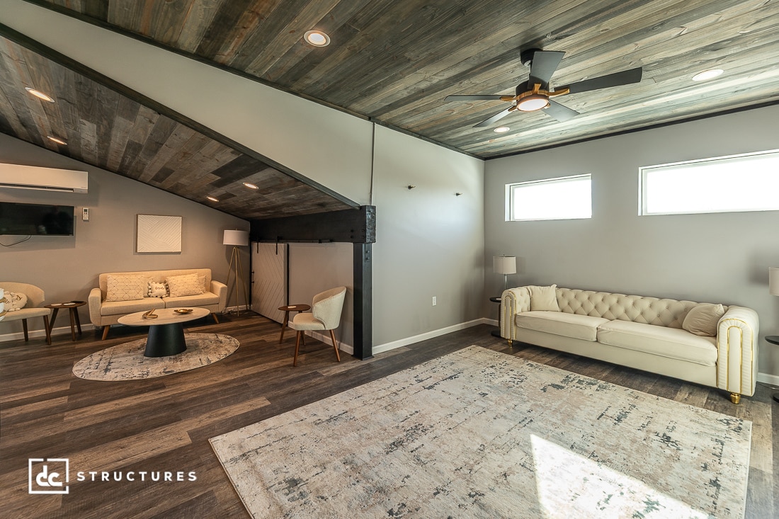 Modern living room with wood-paneled slanted ceiling, ceiling fan, beige sofas, round tables, and sunlight from windows.