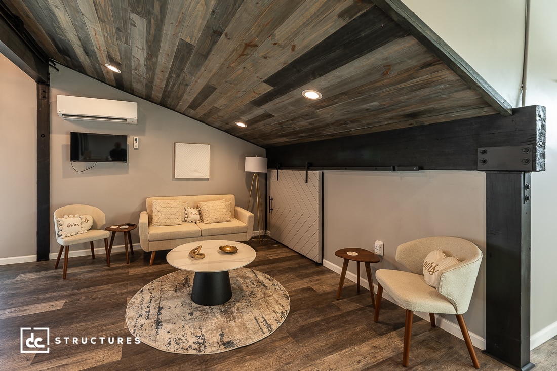 A cozy modern living room with sloped wooden ceiling, beige sofa, armchairs, round rug, coffee table, TV, and white sliding door.
