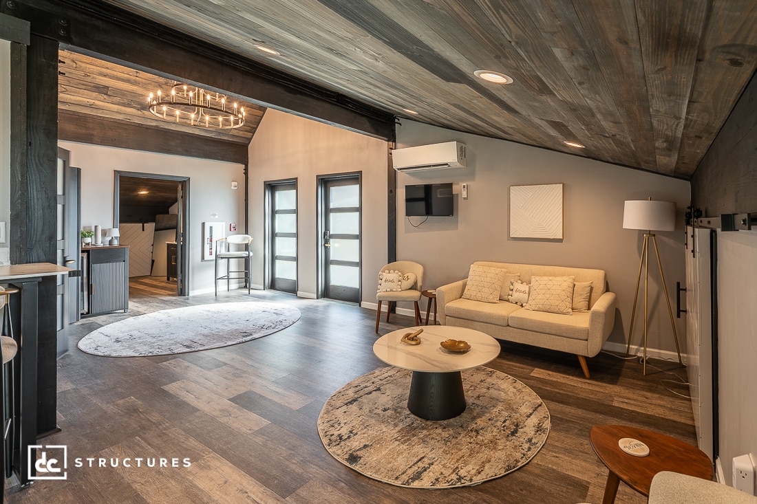 Modern living room with wood floors and ceilings, beige sofa, circular rug, coffee table, TV, large windows, and cozy lighting.
