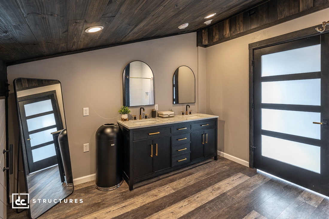 Modern bathroom with wood floors, double-sink vanity with black cabinets and gold handles, two arched mirrors, large floor mirror, black door with frosted glass panels, and recessed lighting.