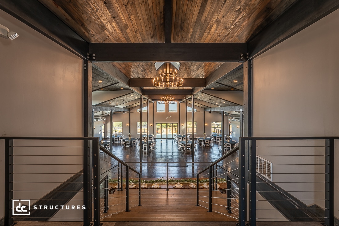 A wide, open event space with wooden ceilings, modern chandeliers, large windows, and set tables. Stairs with metal railings lead down to the main area. The space feels rustic and elegant.