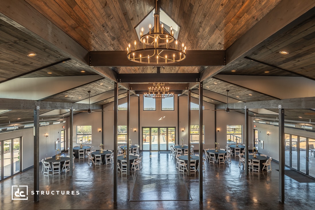 A spacious, modern banquet hall with large windows, wooden ceilings, exposed beams, and elegant chandeliers. Multiple round tables with black and white chairs are arranged neatly on a polished floor.