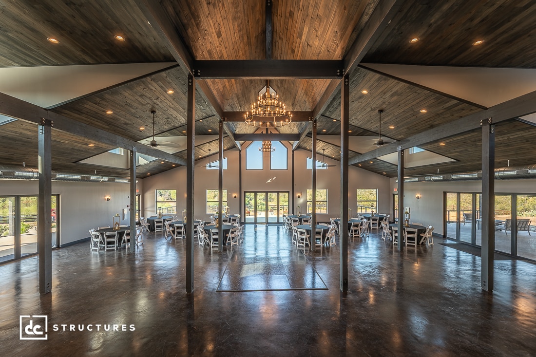 Spacious, elegant event hall with high wooden ceilings, large windows, round tables with white chairs, chandelier, concrete floors.