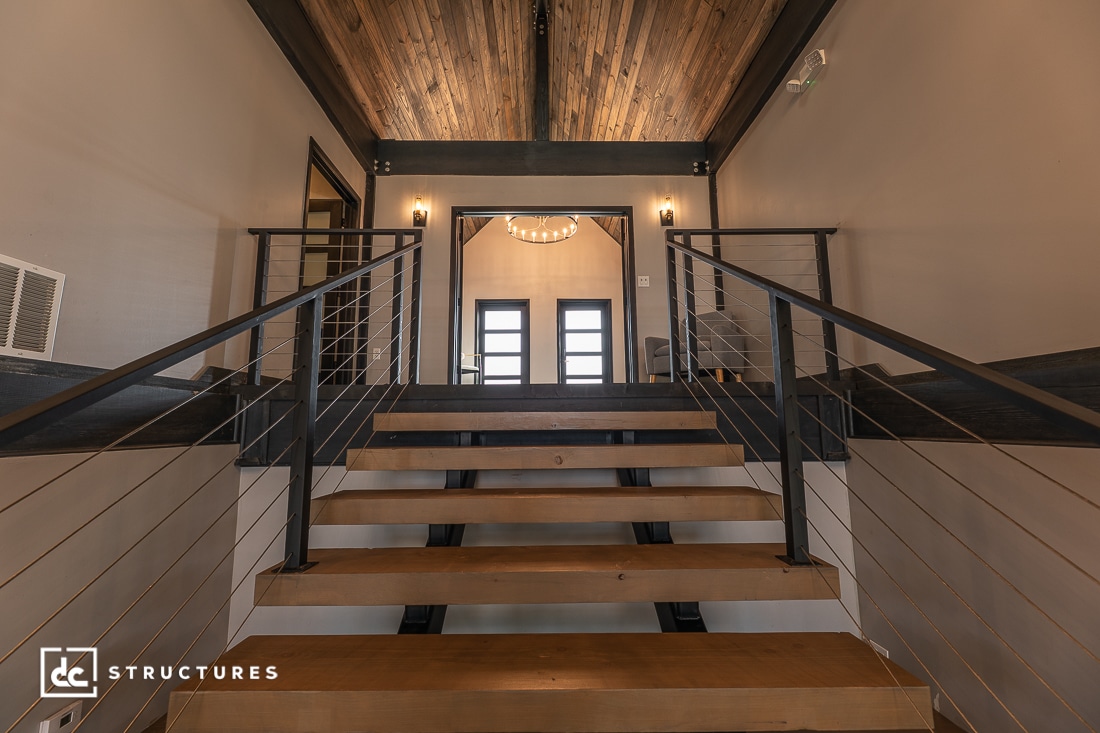 A modern interior with a wooden staircase, metal railings, and vaulted ceiling beams. Double doors and a round chandelier are at the top.