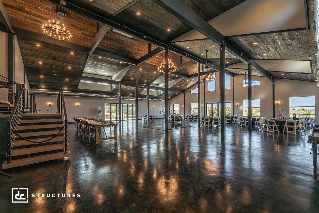 Spacious modern barn event venue with polished concrete floors, high wooden ceilings, large windows, chandeliers, and banquet tables.
