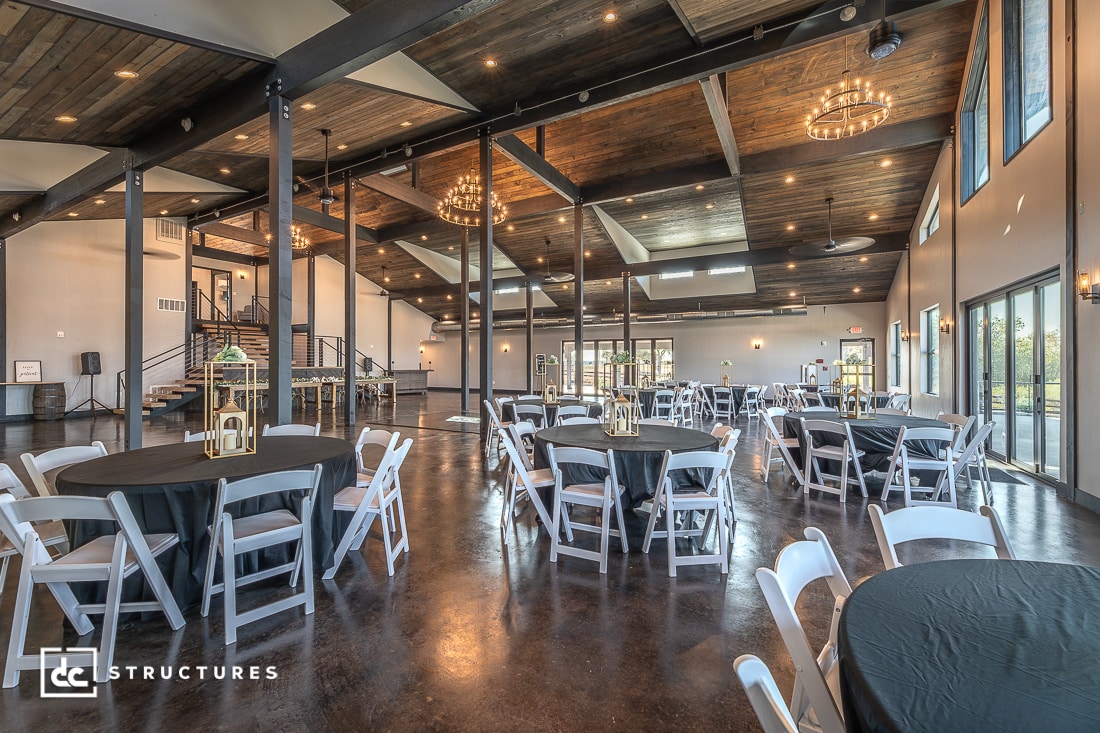 Spacious modern event hall with high wooden ceilings, large windows, and round tables with black tablecloths and white chairs.