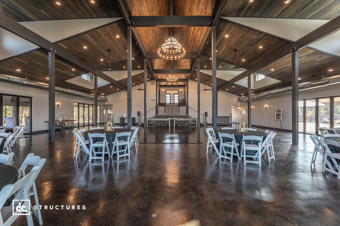 Spacious event hall with high wooden ceilings, large chandeliers, polished concrete floors, round tables, white chairs, and large windows. A raised stage is centered at the far end.