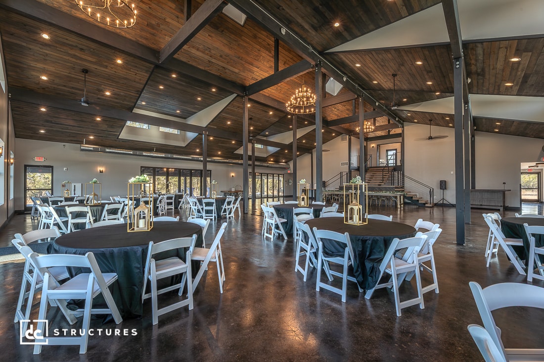 Spacious event hall with high wood ceilings, exposed beams, chandeliers, round tables with black cloths, white chairs, gold lanterns.