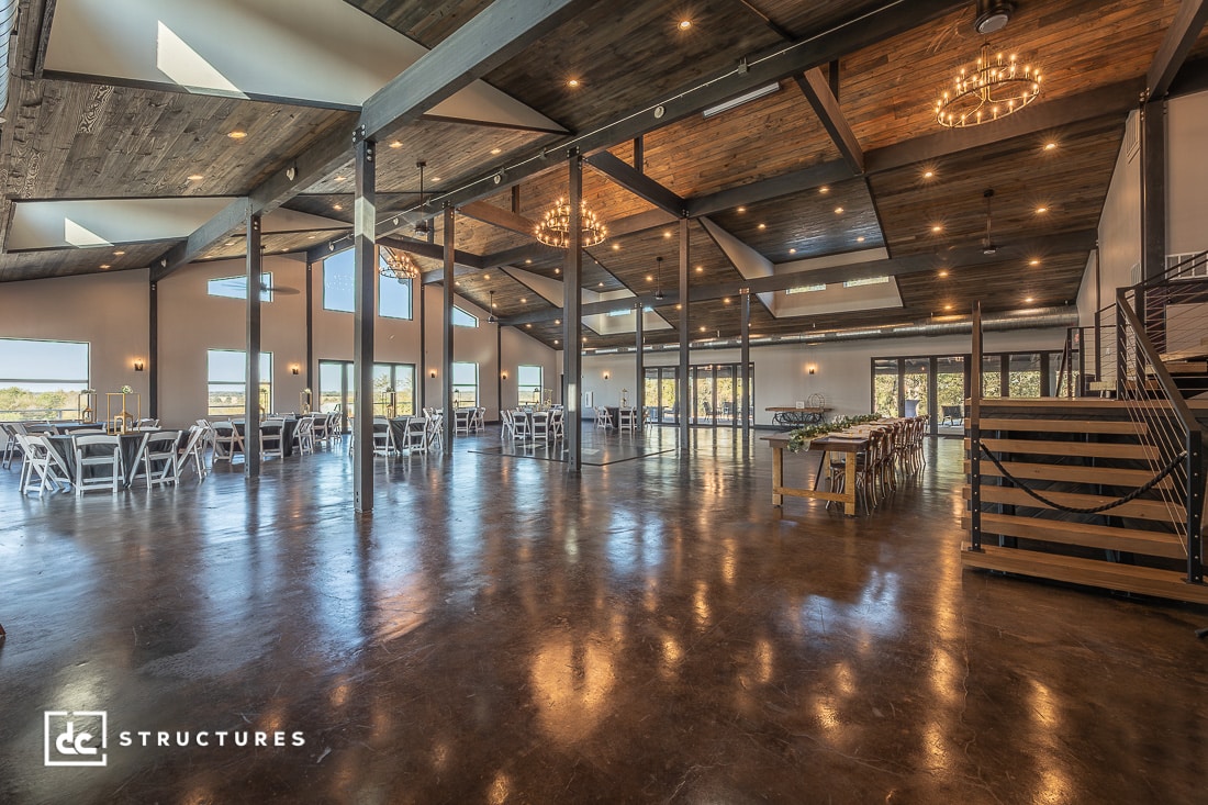 A spacious, modern event hall with polished concrete floors, wooden ceiling, large windows, chandeliers, and rows of white chairs and tables flooded with natural light.