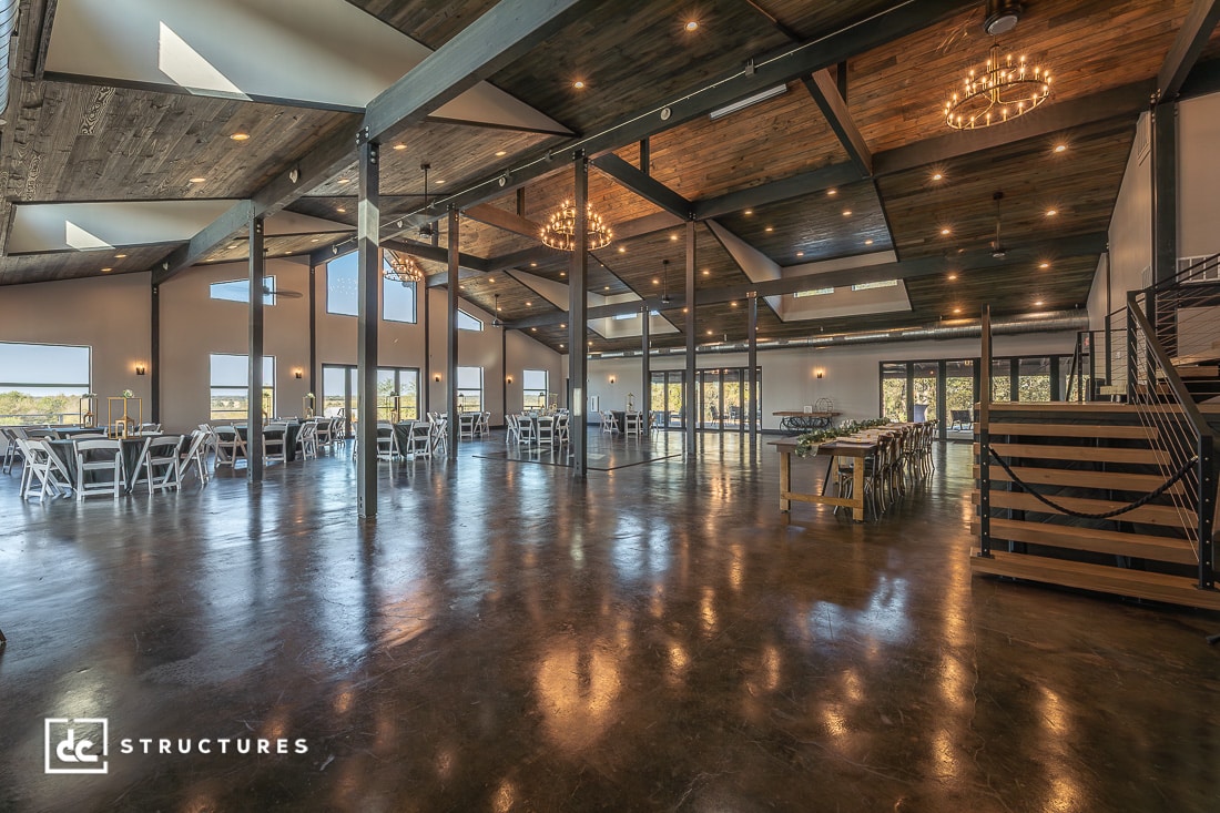Spacious, modern event hall with high wooden ceilings, exposed beams, chandeliers, large windows, and white chairs at round tables.