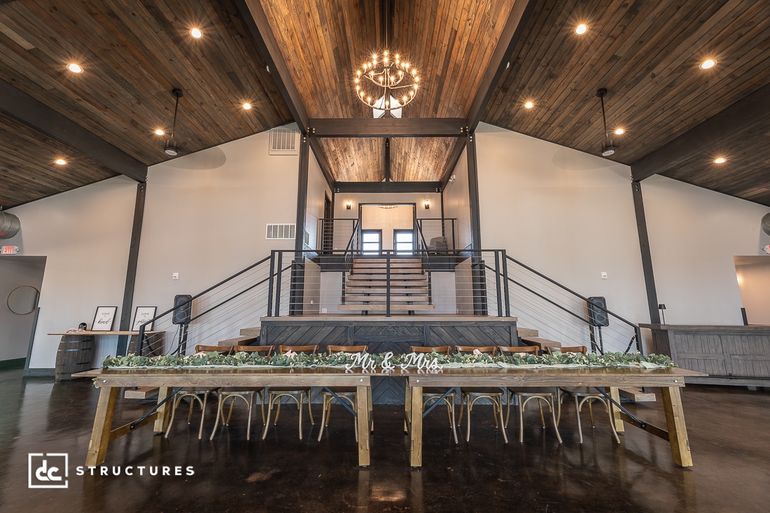 A spacious event hall with a wooden ceiling, a large chandelier, a long decorated table, and a central staircase to an upper level. The room features modern rustic decor.