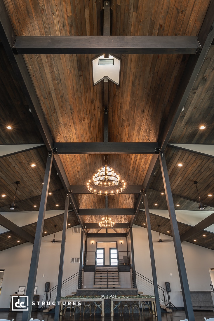 Interior view of a spacious hall with dark wooden vaulted ceilings, exposed beams, chandeliers, and a staircase to a raised area.