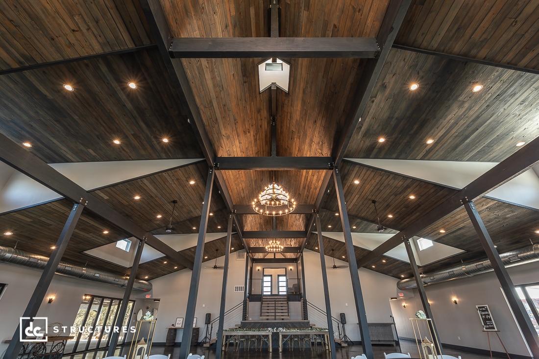 A spacious modern event hall with exposed wooden beam ceilings, hanging chandeliers, large windows, and a central staircase.