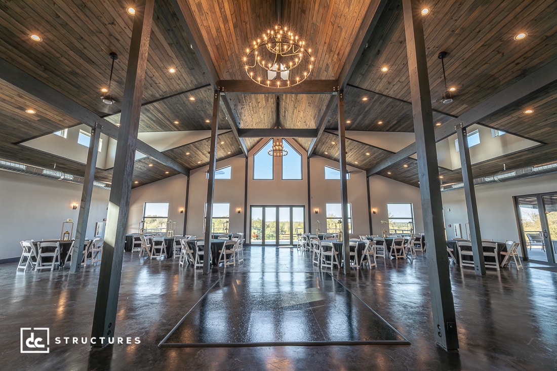 Spacious modern event hall with exposed beams, vaulted wood ceiling, large windows, chandeliers, dark floor, white tables and chairs.