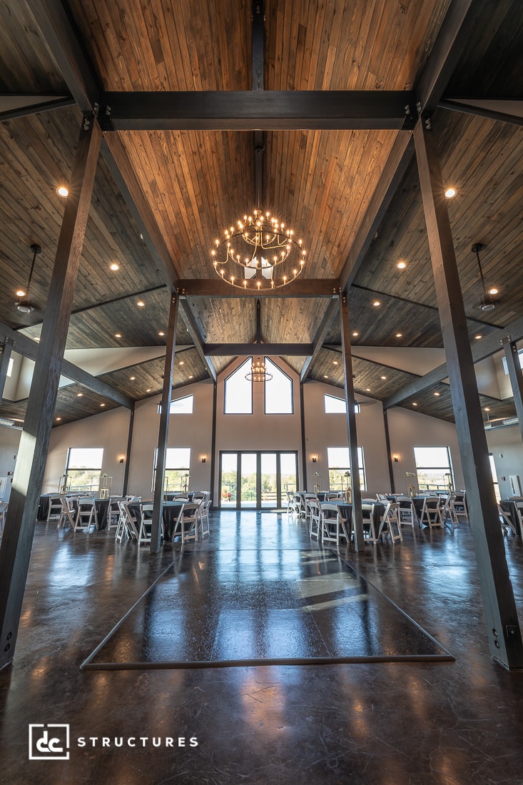 Spacious modern event hall with a high wooden ceiling, large windows, chandeliers, polished floor, and rows of white chairs and tables.