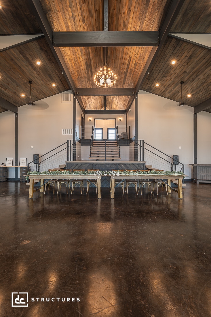 A spacious event hall with high wooden ceilings, a large chandelier, and a long rustic dining table with chairs. Stairs lead to a raised platform at the back, and the floor is polished concrete.