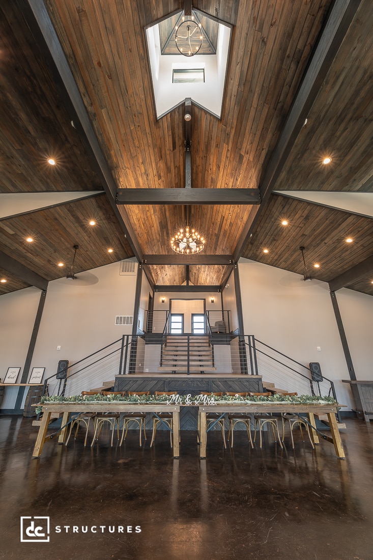 A spacious event hall with high wooden ceilings, a grand chandelier, a banquet table with greenery, and a balcony staircase.