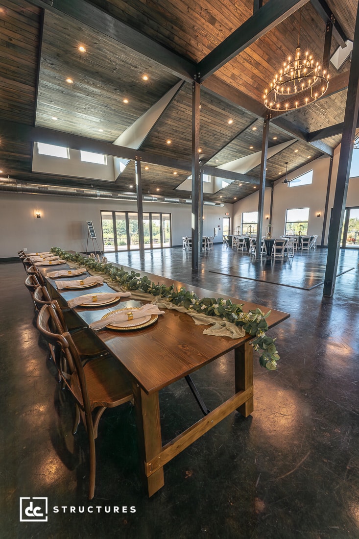A spacious, modern event hall with high wooden ceilings and large windows. Long wooden table with greenery decor in the foreground.
