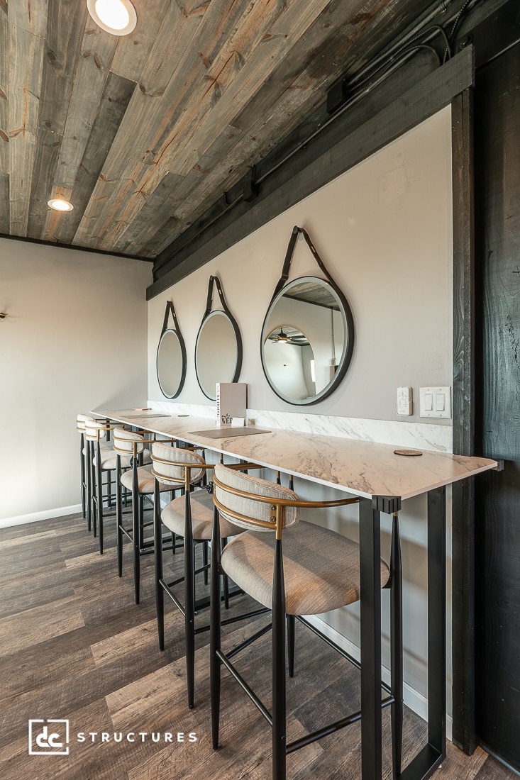 A modern bar area with three high marble tables and upholstered stools, round mirrors on a gray wall, and wood ceiling and floor.