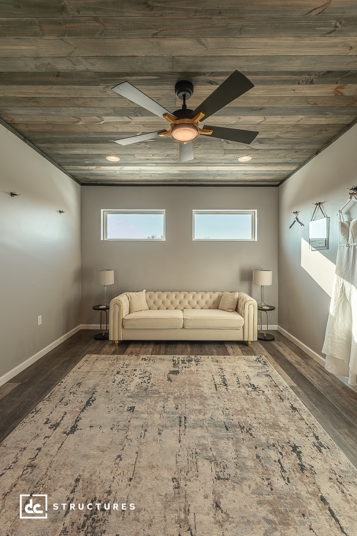 A modern room with a beige tufted sofa, gray wall, two windows, side tables with lamps, ceiling fan, rug, and bridal dresses.