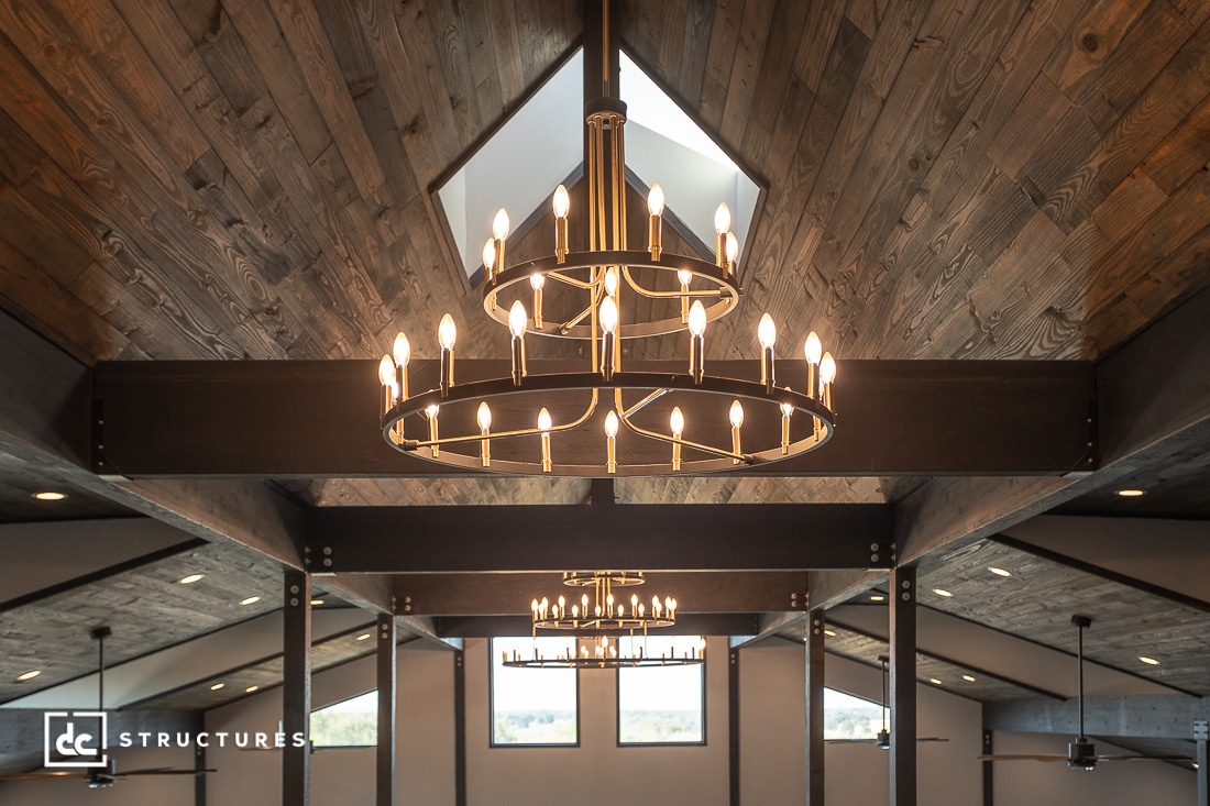 A large modern chandelier with multiple candle-shaped bulbs hangs from a high wood-paneled vaulted ceiling in a spacious room.