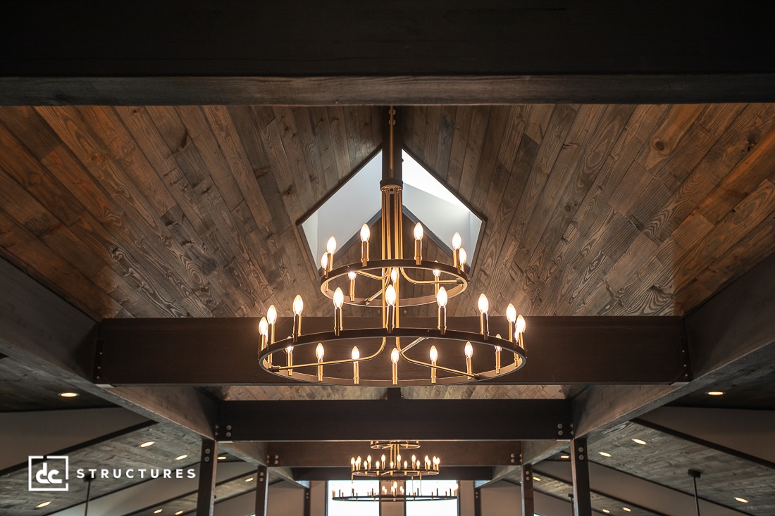 Large round chandelier with lit candle-style bulbs hangs from a vaulted wooden ceiling with exposed beams. Natural light enters through upper windows.