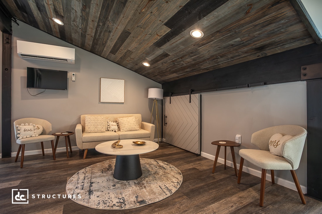 A modern loft living room with a sloped wooden ceiling, beige sofa, two armchairs, round rug, coffee table, TV, and sliding barn door. Cozy, rustic feel.
