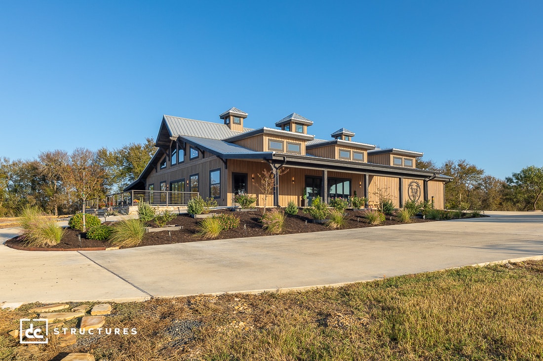 A modern barn-style building with a metal roof, large windows, and landscaped gardens, set against a clear blue sky and trees.