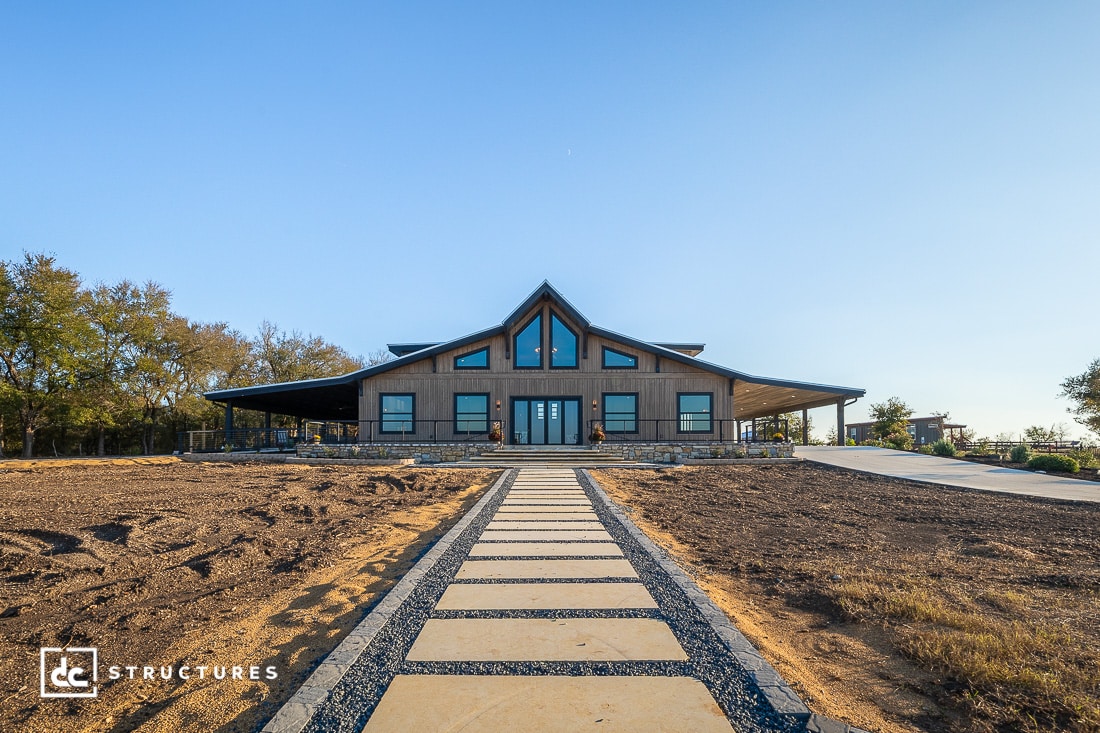 A modern barn-style home with large windows and a wide wraparound porch sits at the end of a stone walkway, surrounded by dry grass and trees under a clear blue sky.